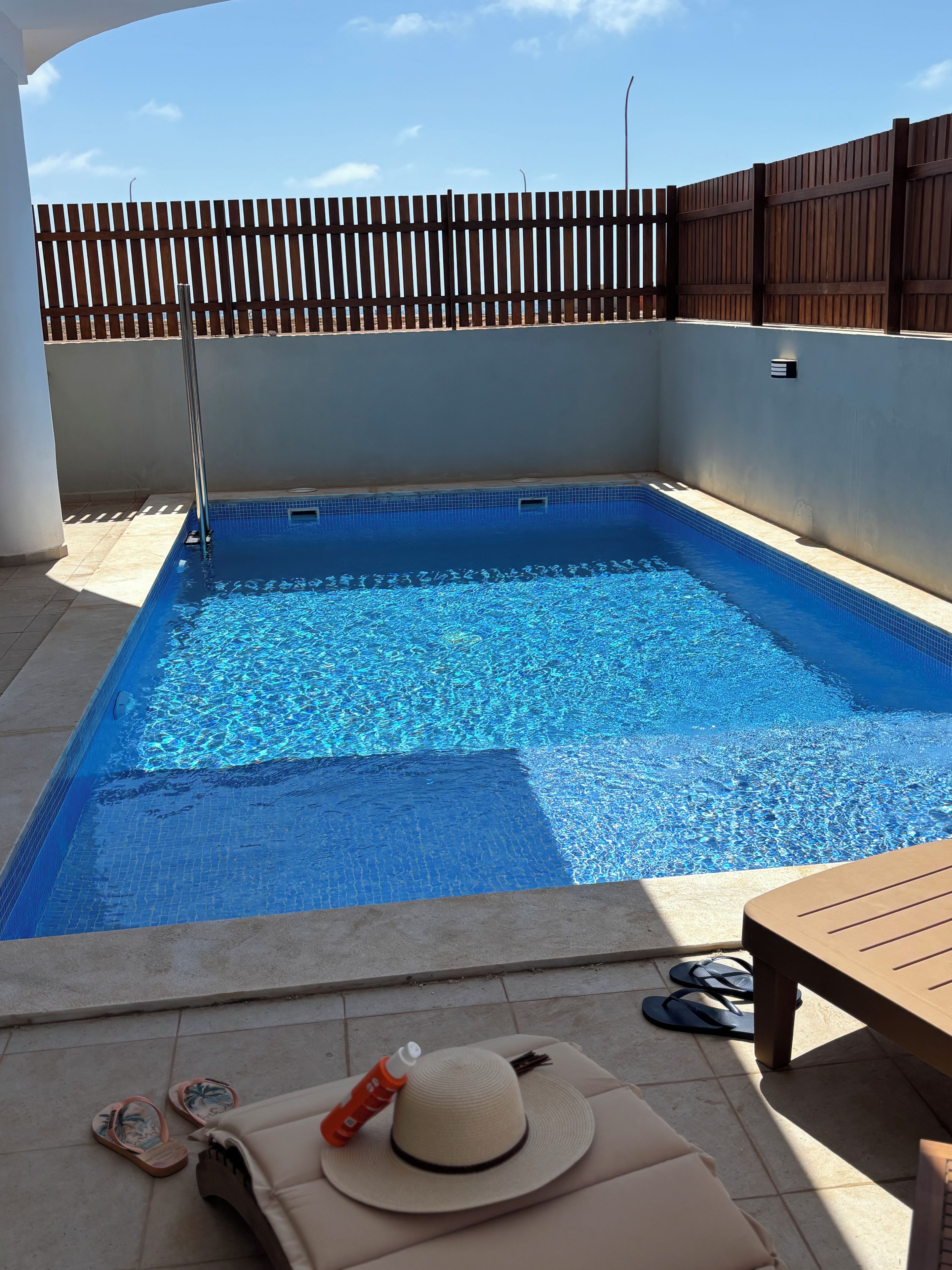 A swimming pool with a wooden fence, a sun lounger, and a hat on the lounger.