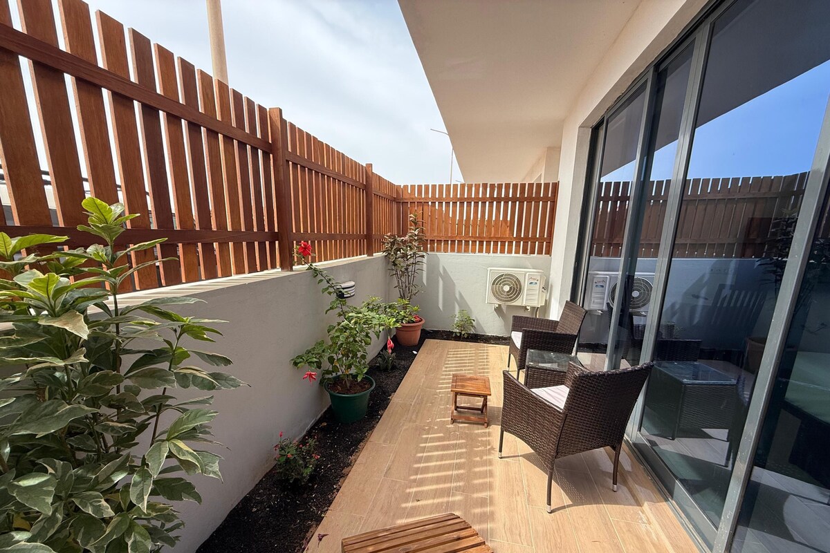 A balcony with a wooden floor, potted plants, a small table, and wicker chairs.