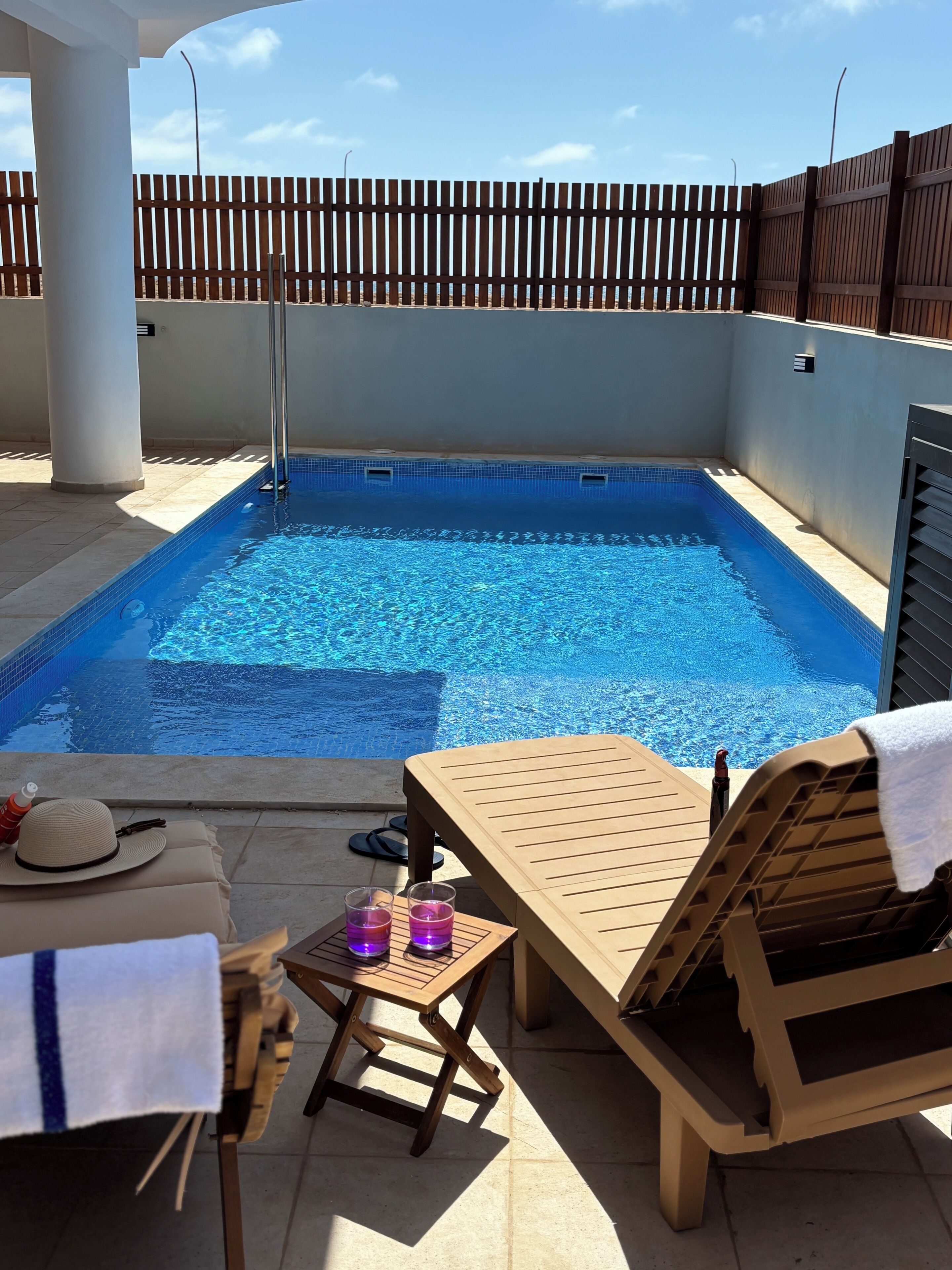 A swimming pool with a wooden deck, lounge chairs, and a table with drinks and a hat.