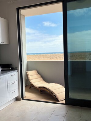 A balcony with a beach view, a cushioned lounge chair, and a kitchen area with cabinets and a countertop.