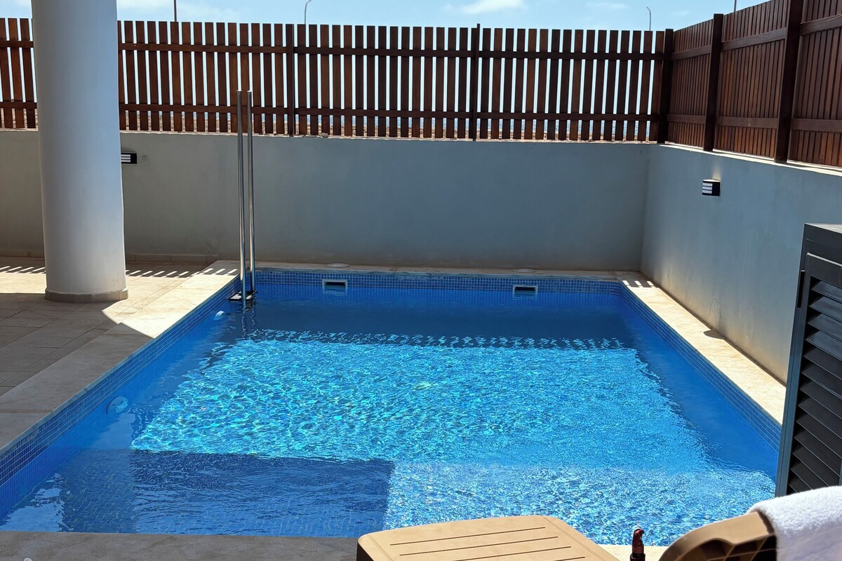 A swimming pool with a wooden deck, lounge chairs, and a table with drinks and a hat.