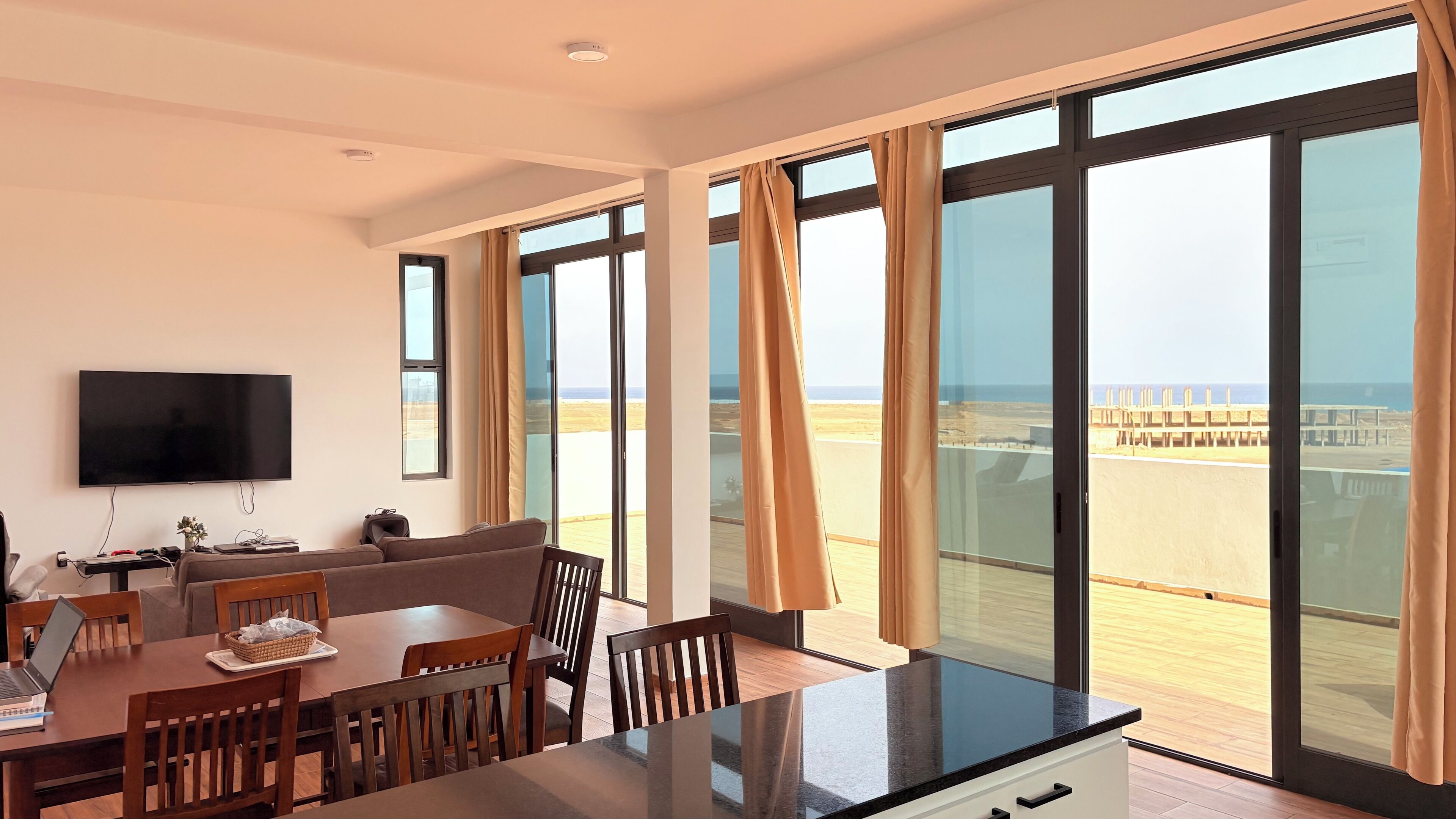 A modern hotel room with a dining area, a flat-screen TV, and large windows offering a view of the beach.