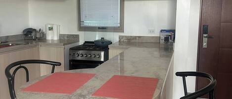 A modern kitchen with a central island, two bar stools, a stove, and a window with blinds.