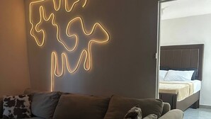 A modern living room with a sectional sofa, a neon light art piece on the wall, and a glimpse of a bedroom with a bed and nightstand. - Modern Luxury Condo With Pool Access in Kingston, Jamaica (Kingston)