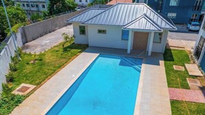 A rectangular swimming pool with a tiled deck, surrounded by a small house with a metal roof and a paved driveway. - Modern Luxury Condo With Pool Access in Kingston, Jamaica (Kingston)