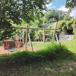 Un parque infantil con columpios y estructuras para escalar, rodeado de follaje y una valla de madera.