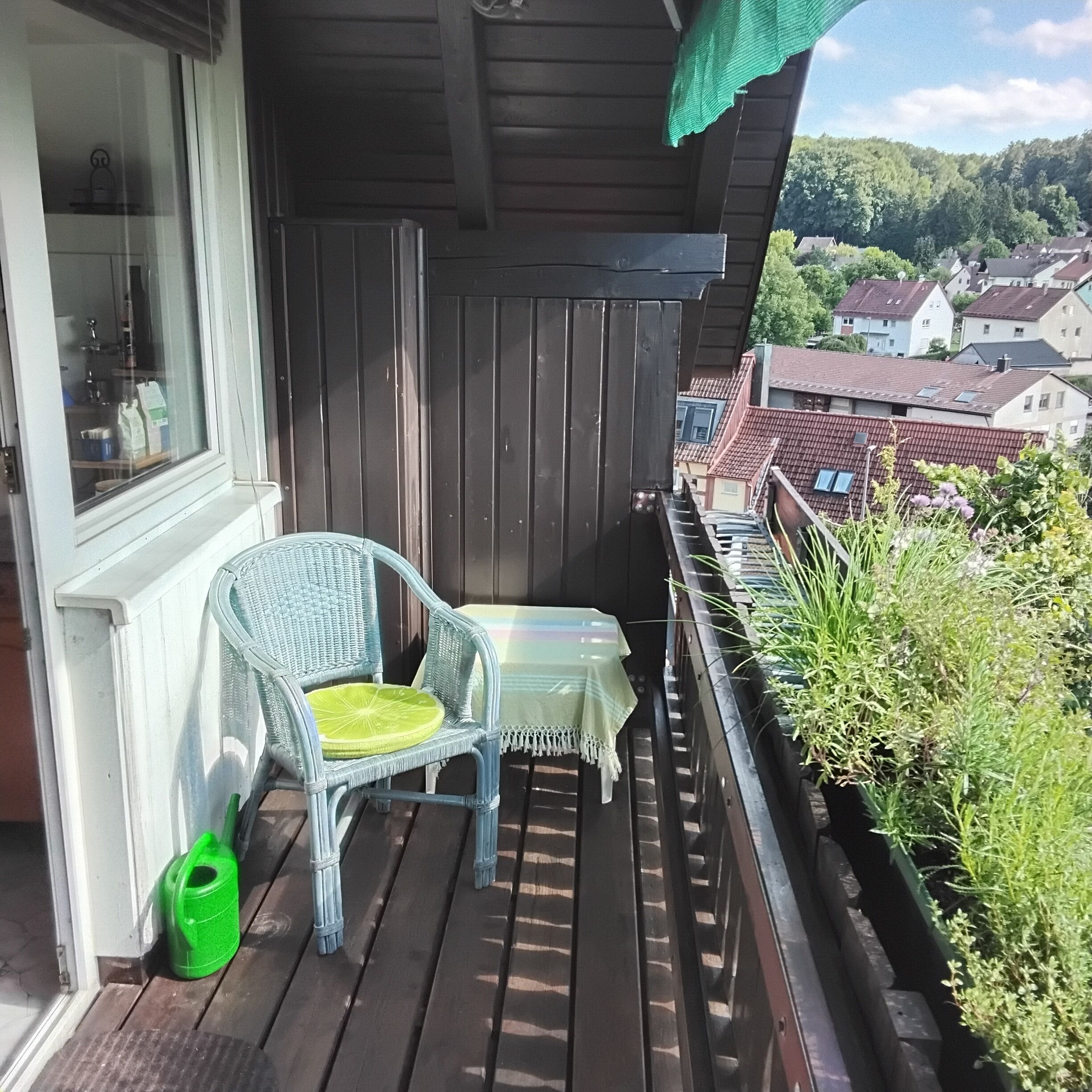 Een houten balkon met een rieten stoel, een tafeltje en een groene plantenbak.