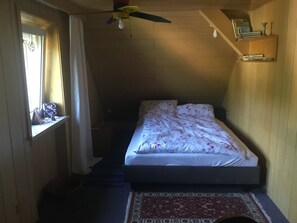 Ein Schlafzimmer mit einem Bett, einem Fenster mit Vorhängen, einem Deckenventilator und einem kleinen Regal mit Büchern und einer Vase.