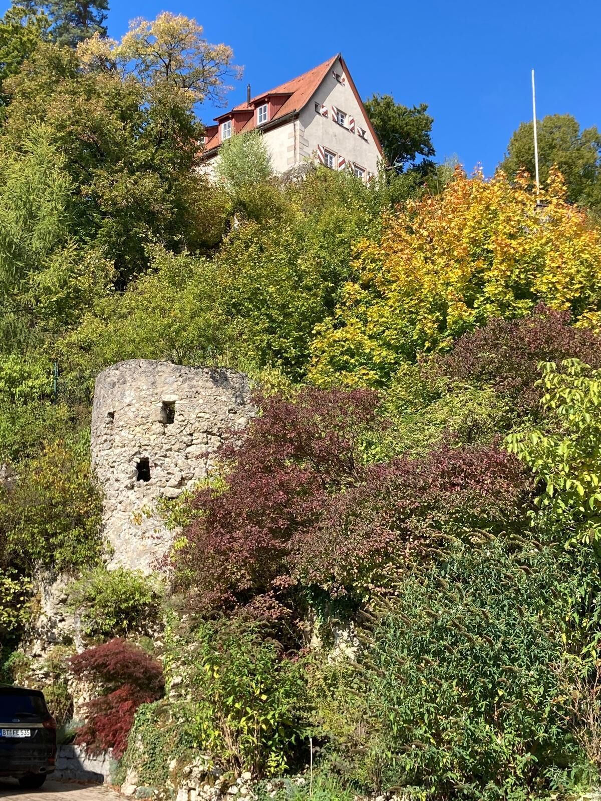 En historisk bygning med et stenhus og en struktur med rødt tag, omgivet af frodigt grønt.