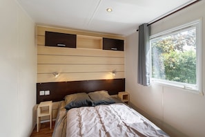 Une chambre à coucher comprenant un lit, des tables de chevet, une tête de lit dotée d’étagères intégrées, une fenêtre avec des rideaux et une vue sur la verdure à l’extérieur.