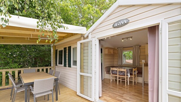 Een overdekt buitengebied met een tafel, stoelen, een houten terras en de ingang van een gebouw.