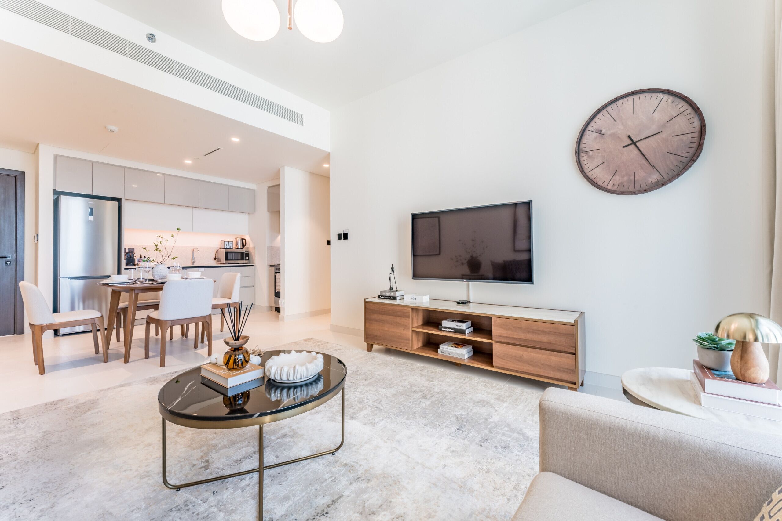 Een moderne woonkamer met een flatscreen televisie, een houten tv-meubel, een ronde klok en een eethoek met een tafel en stoelen.