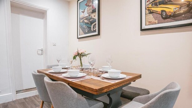 A dining area with a wooden table set for four, grey upholstered chairs, and framed artwork on the wall. - Gloucester-5 Bed Home for Work & Leisure (Gloucester)