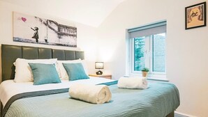 A hotel room with a bed, pillows, a nightstand, a lamp, a window with a view, and artwork on the wall. - Gloucester-5 Bed Home for Work & Leisure (Gloucester)