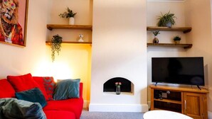 A living room with a red sofa, a flat-screen TV, wooden shelves with plants, and a fireplace. - Gloucester-5 Bed Home for Work & Leisure (Gloucester)
