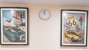 A dining table set for four with white plates, glasses, and cutlery. Above the table are two framed posters, one of a motel and the other of Route 66. A clock is mounted on the wall above the table. - Gloucester-5 Bed Home for Work & Leisure (Gloucester)
