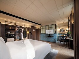 A hotel room with a large bed, a desk with a chair, a minibar, a wardrobe, and a bathroom with a mirror.