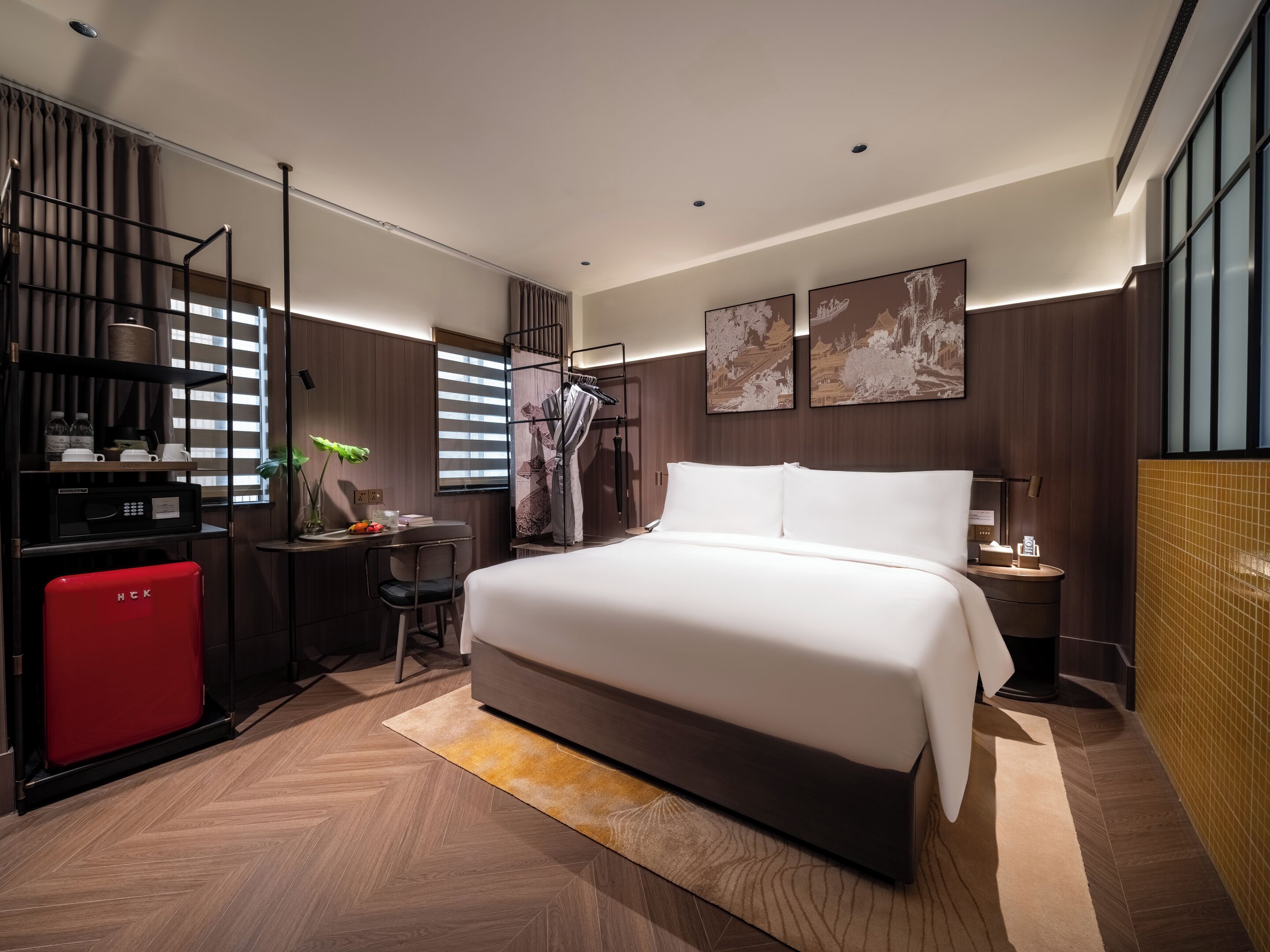 A modern hotel room with a large bed, a desk, a red minibar, and artwork on the walls.