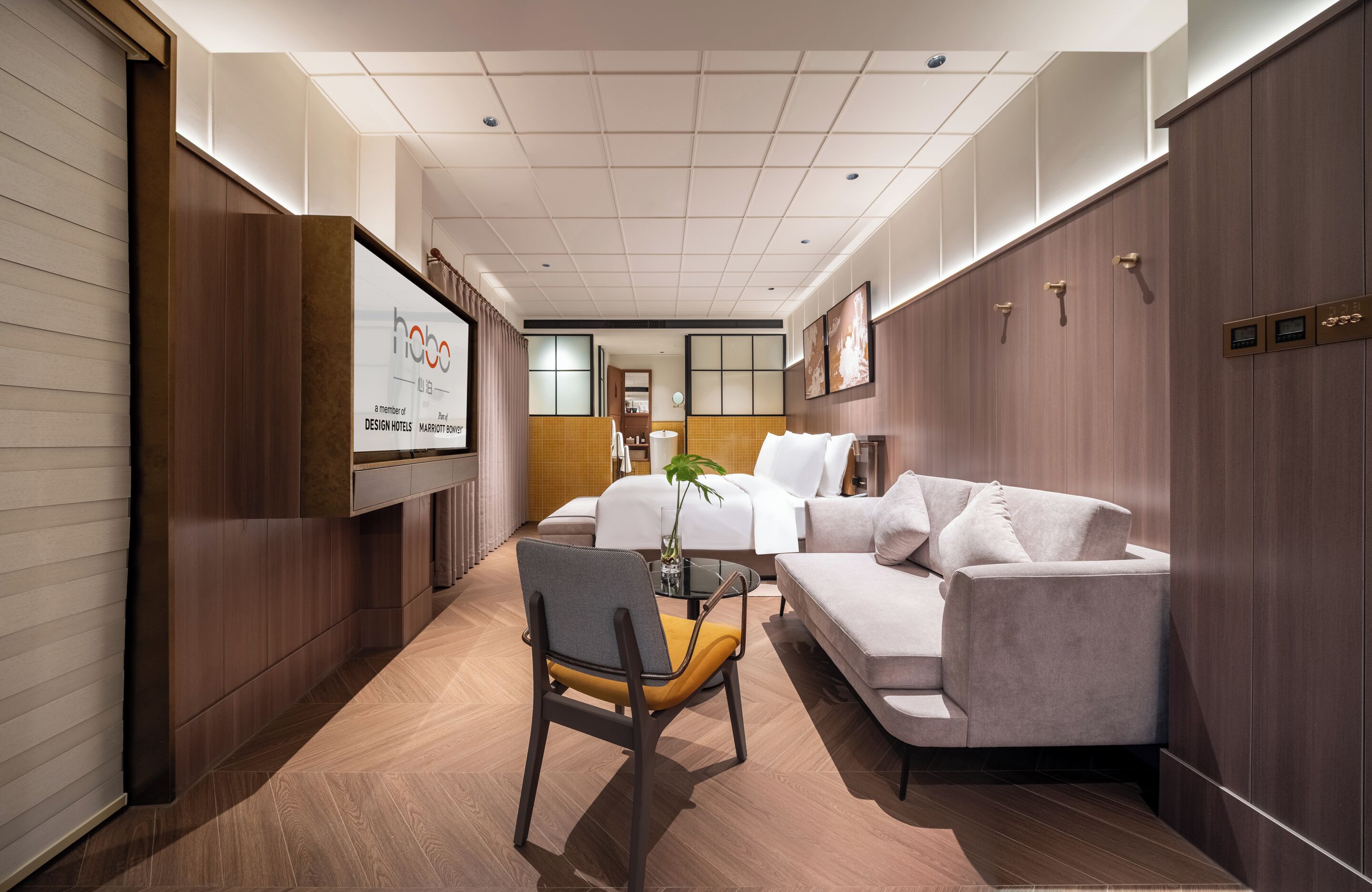 Ein Hotelzimmer mit einer Couch, einem Sessel, einem kleinen Tisch, einem Bett und einem Fernseher, der den Namen des Hotels anzeigt.