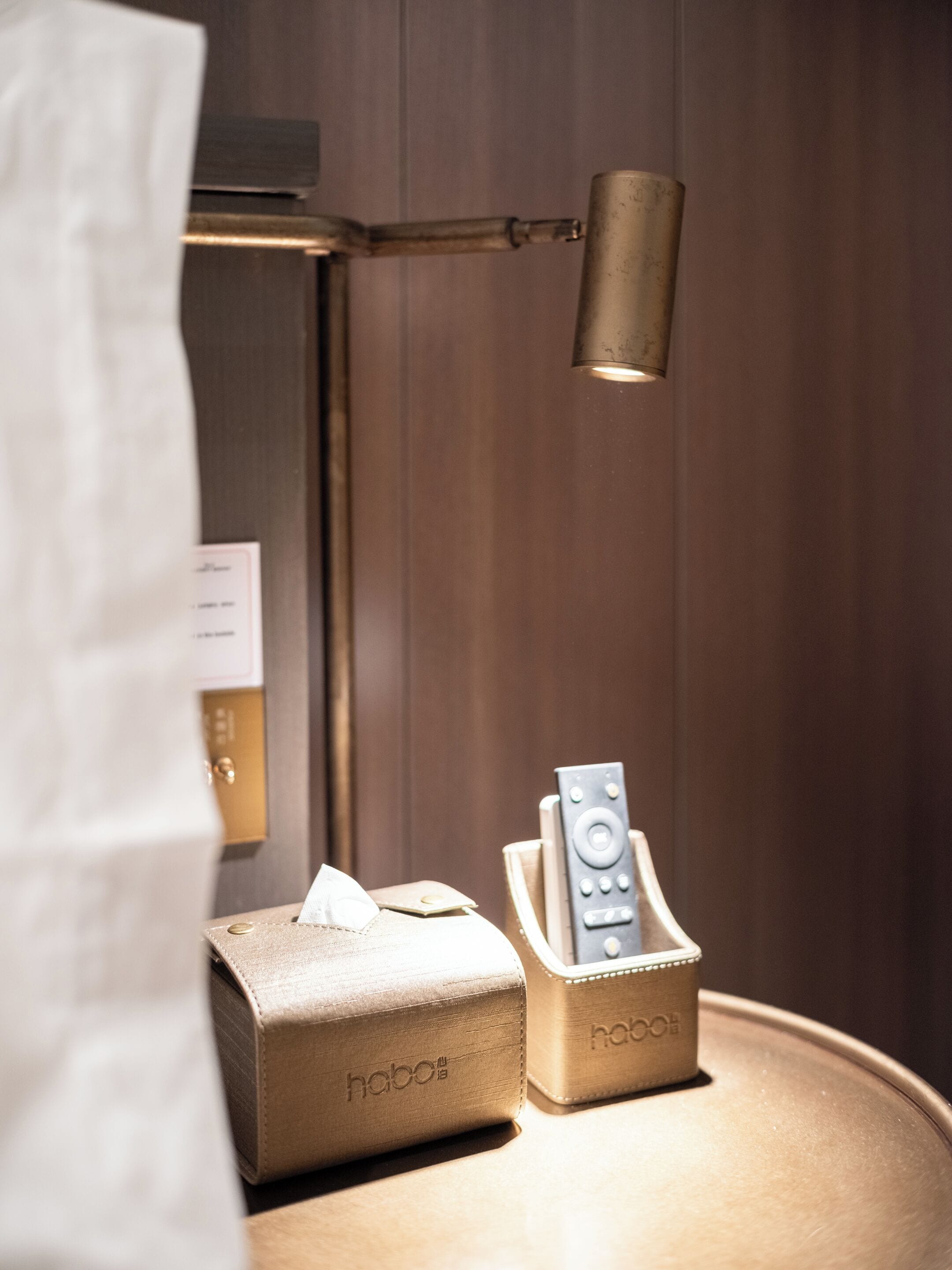 A bedside table with a remote control and a tissue box, a lamp, and a curtain partially visible.