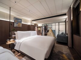 A hotel room with a large bed, a seating area with a chair, a wardrobe, a bedside table with a lamp, and a painting on the wall.