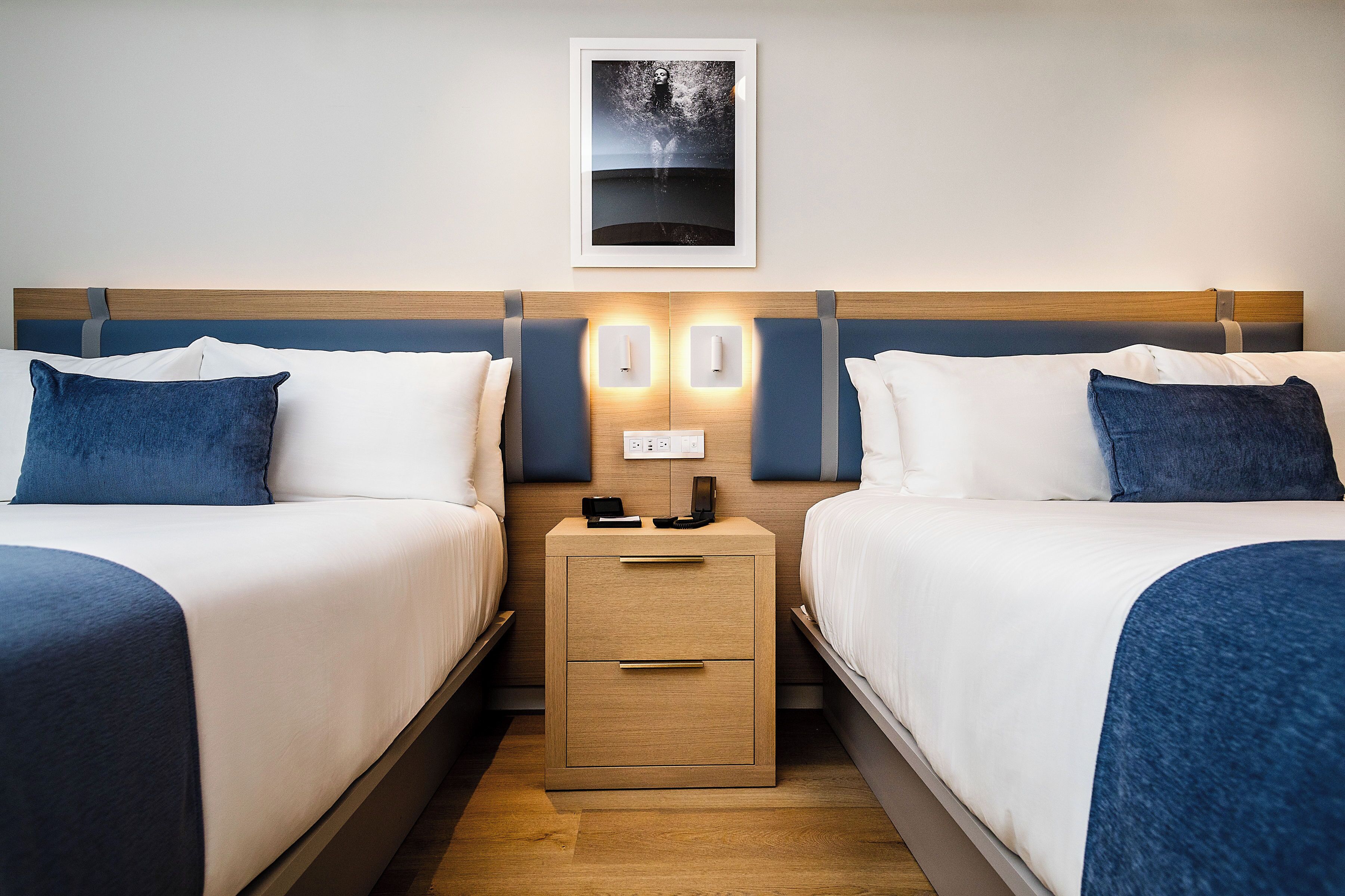 Une chambre d’hôtel avec deux lits, une table de chevet avec un téléphone et un chargeur, et un tableau encadré au mur.