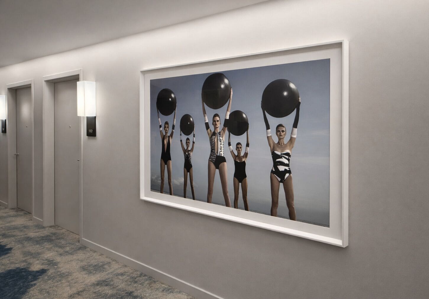 A hallway with a large framed picture of five individuals holding black balloons, a carpeted floor, and a door with a number sign.