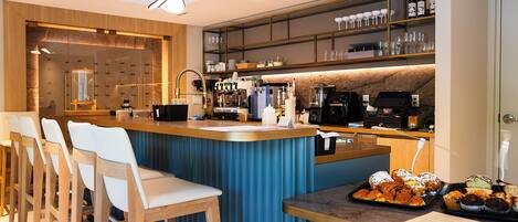 Un espace bar moderne, doté de tabourets, d’une machine à café et d’un assortiment de viennoiseries.