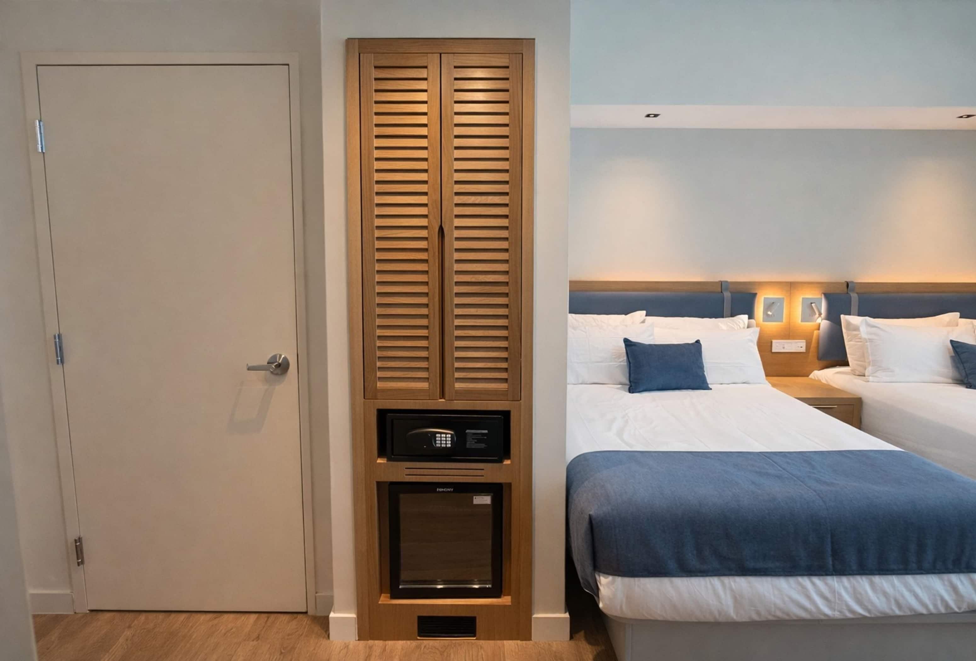 Ein Hotelzimmer mit zwei Betten, einem Kleiderschrank, einer Minibar und einem Nachttisch mit Lampe.
