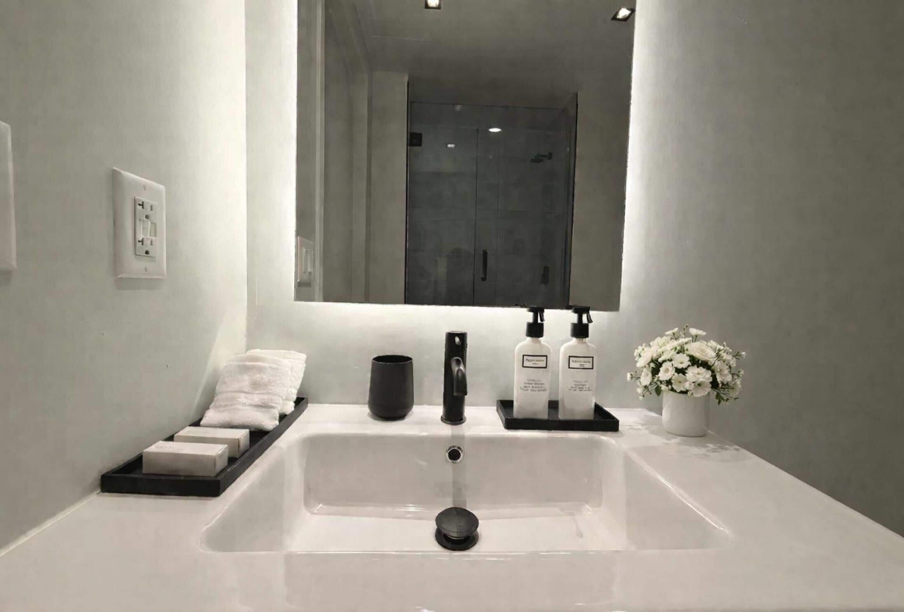 Un baño con un espejo grande, un lavamanos blanco, un grifo negro y un dispensador de jabón negro.