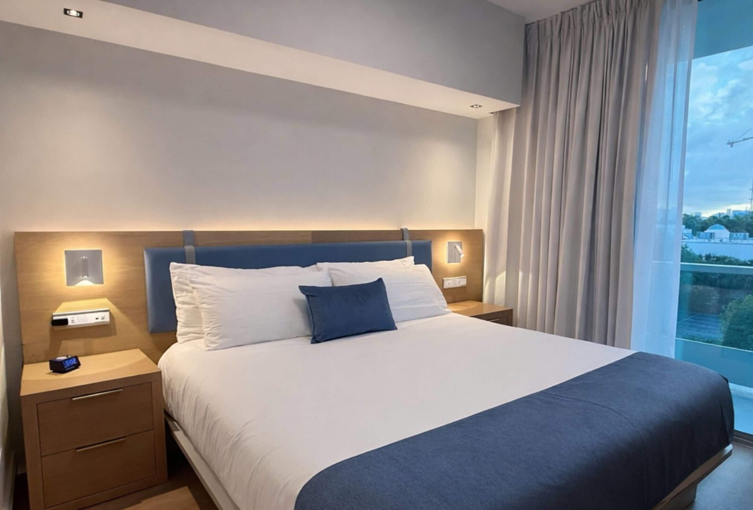 A hotel room with a bed, bedside tables, a nightstand, a window with curtains, and a view of buildings outside.