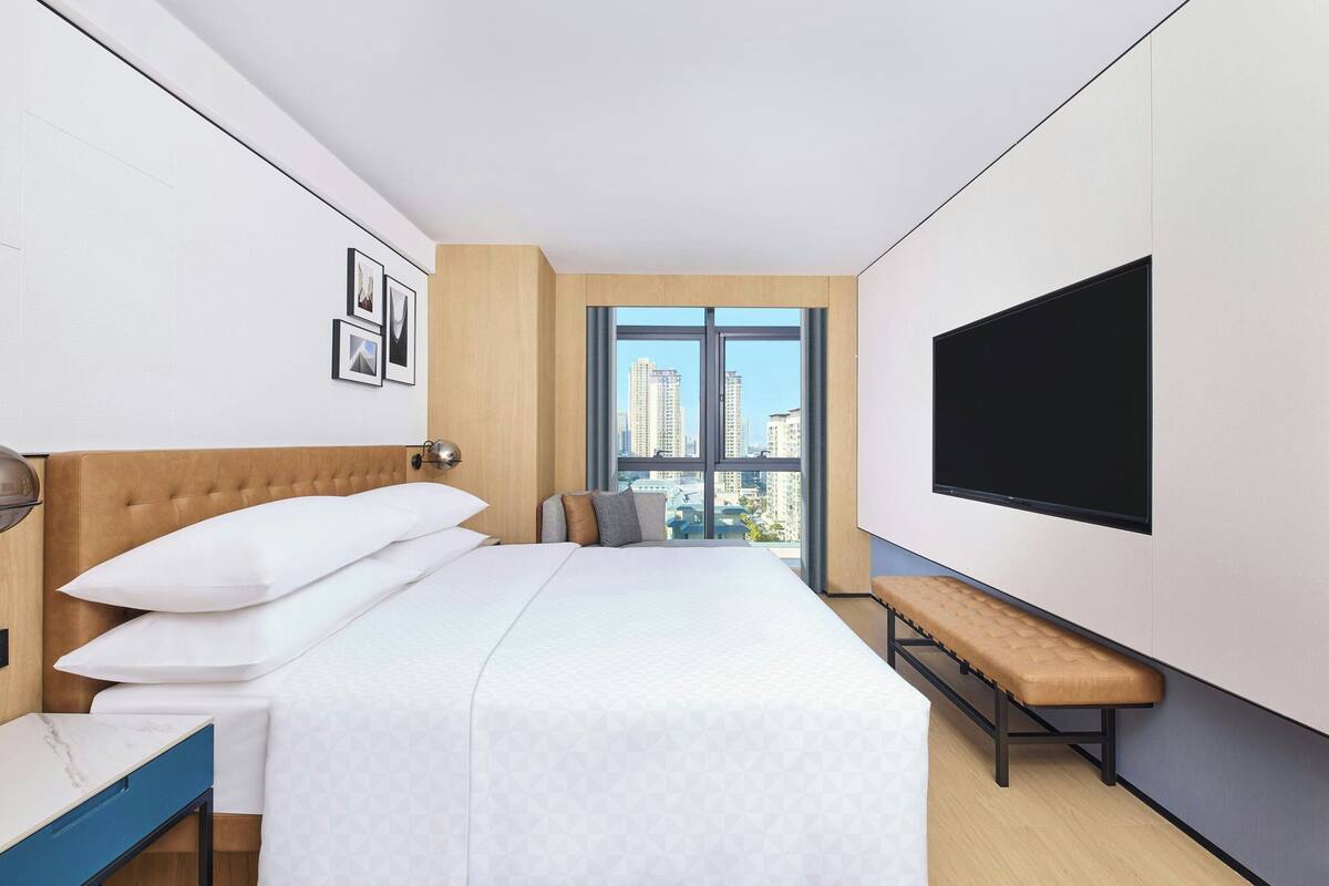 Une chambre d’hôtel moderne avec un grand lit, un banc, une télévision et une vue sur la ville.