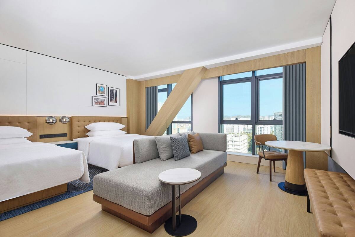 Une chambre d’hôtel moderne avec un canapé-lit, une table ronde et une vue sur la ville grâce à de grandes fenêtres.