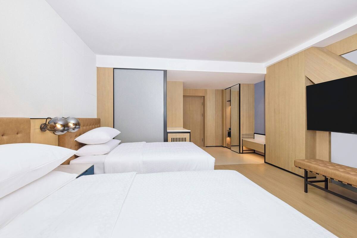 Une chambre d’hôtel moderne avec deux lits, un banc, une télévision et un grand miroir.