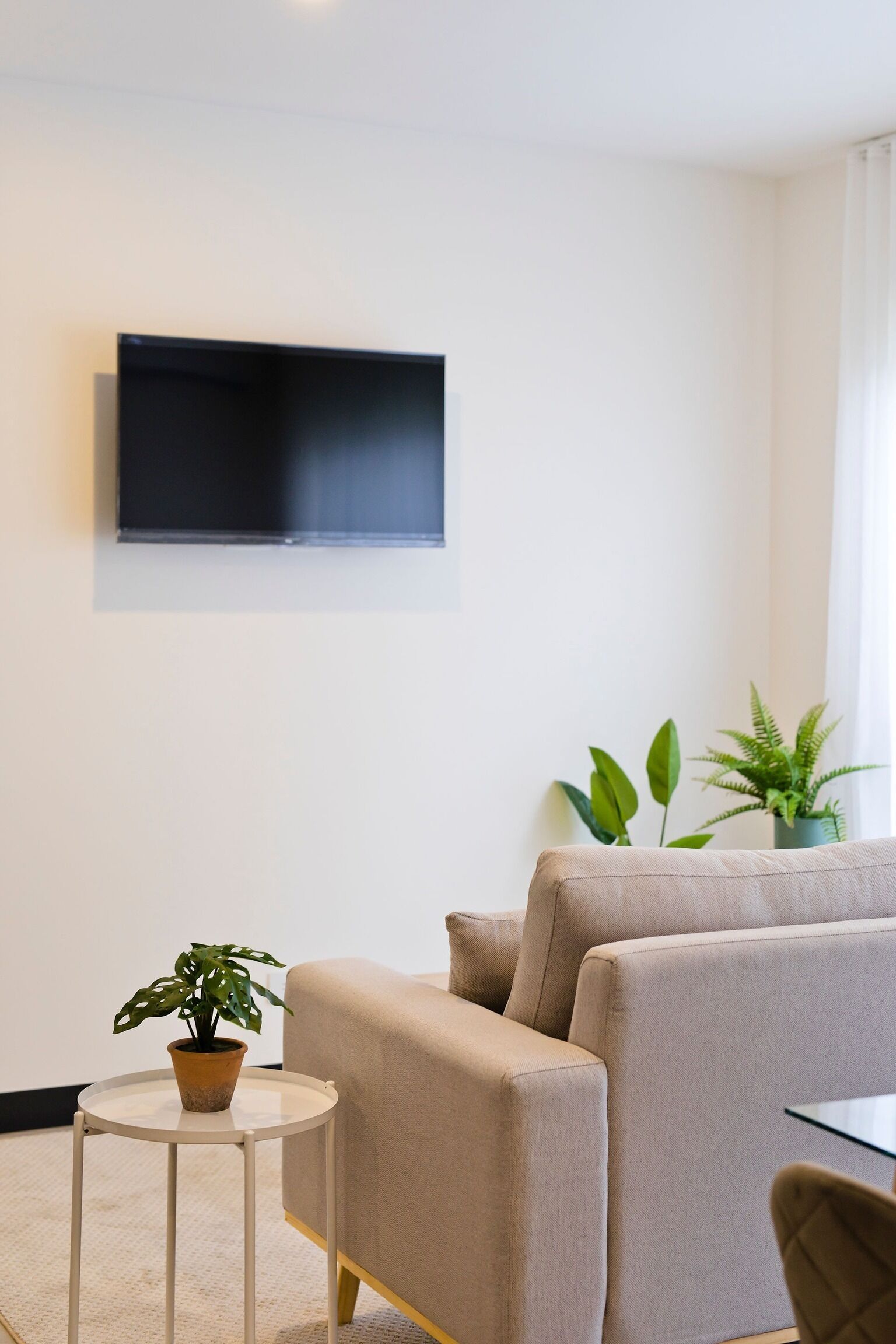 Un salon moderne avec un téléviseur à écran plat fixé au mur, un canapé beige, une petite table ronde ornée d’un pot de fleurs et une autre plante dans un coin.