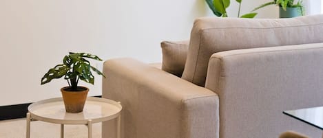 Una sala de estar moderna con un televisor de pantalla plana montado en la pared, un sofá color beige, una mesita redonda con una planta en maceta y otra planta en la esquina.