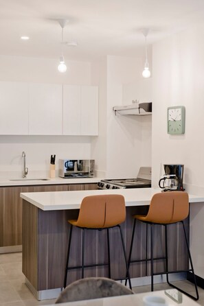 A modern kitchen with white cabinets, a built-in stove, and two bar stools. - Condominio Novatriana - SJO Airport (Alajuela)