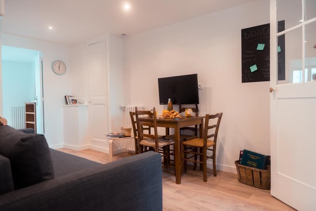 A modern living room with a dark grey sofa, a wooden dining table with chairs, a flat-screen TV mounted on the wall, and a small bookshelf.