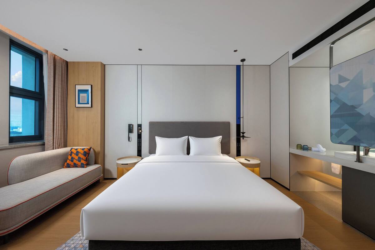 A modern hotel room with a large bed, a sofa, a desk, and a view of the city.