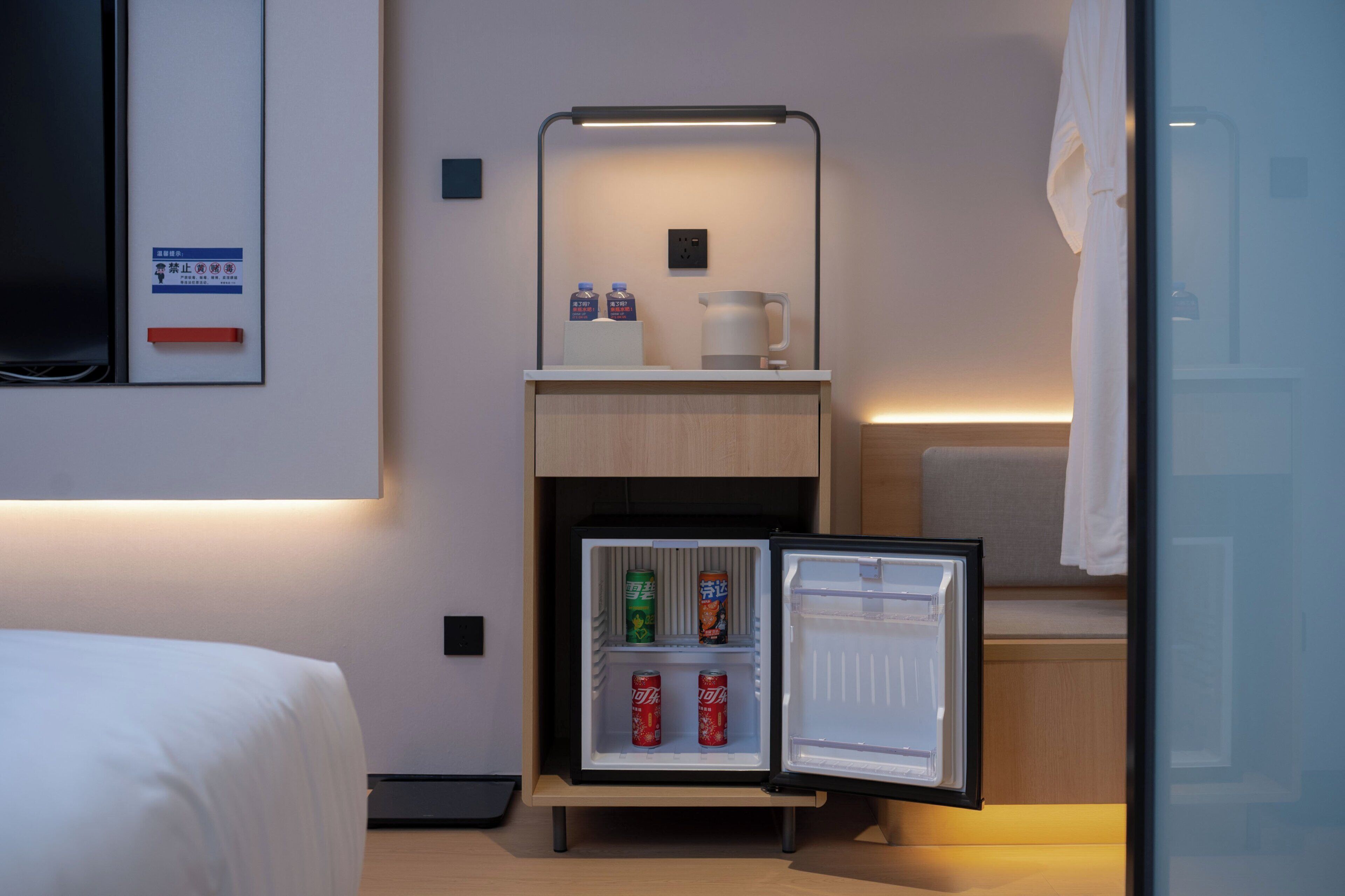 A compact hotel room with a bed, a small refrigerator with beverages, a coffee maker, and a wall-mounted TV.