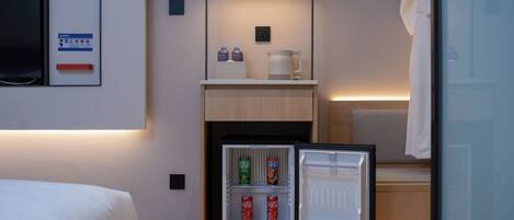A compact hotel room with a bed, a small refrigerator with beverages, a coffee maker, and a wall-mounted TV.