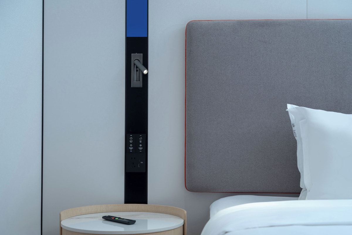 A modern hotel room with a bed, a bedside table with a remote control, a wall-mounted control panel, and a headboard.