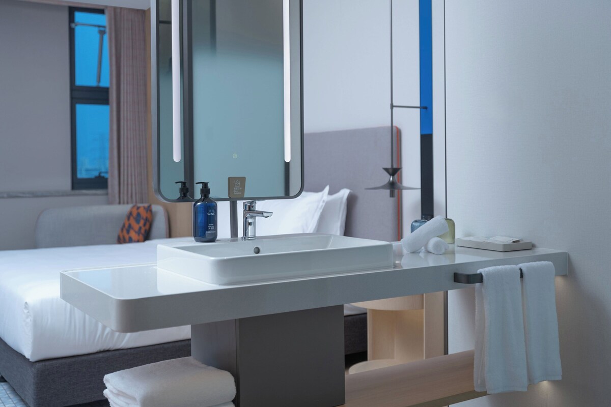 Habitación de hotel moderna con un amplio tocador de baño, cama con ropa blanca, un espejo grande y vistas a la ciudad.