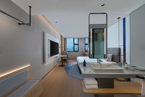 Una habitación de hotel moderna con una cama grande, una mesita de noche, un televisor integrado, un espejo amplio y un baño con lavamanos y espejo.