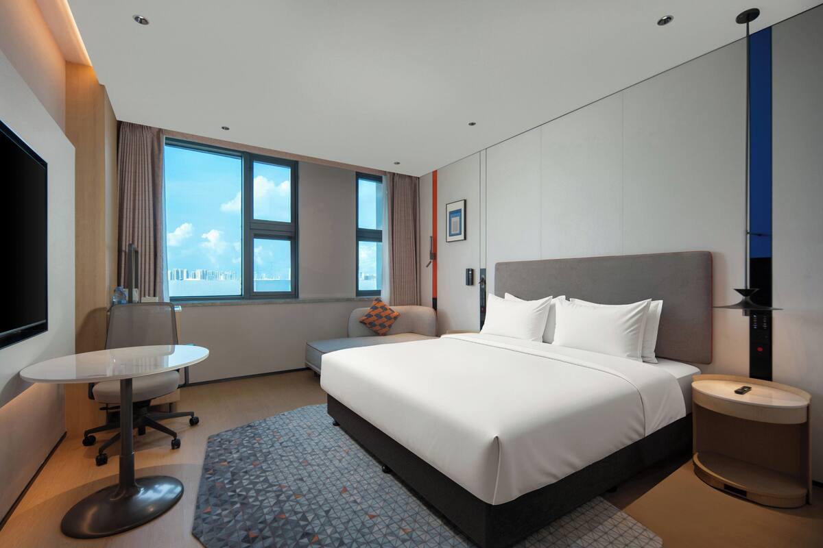 A modern hotel room with a large bed, a desk, a chair, a small table, and a bedside table.