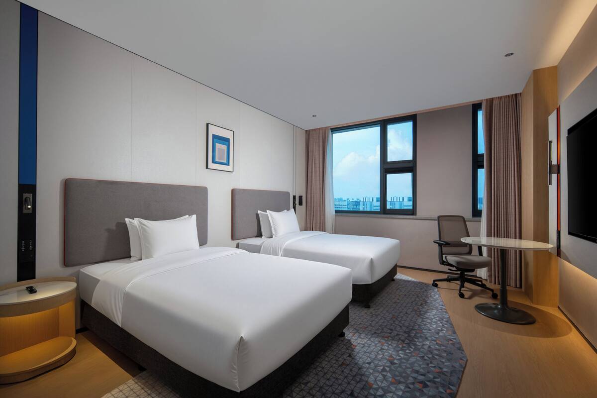 A hotel room with two beds, a desk, a chair, a TV, and a window with a city view.