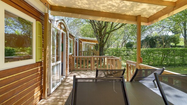Un espacio exterior cubierto con barandillas de madera y una pérgola, que da paso a un jardín con árboles y un seto.