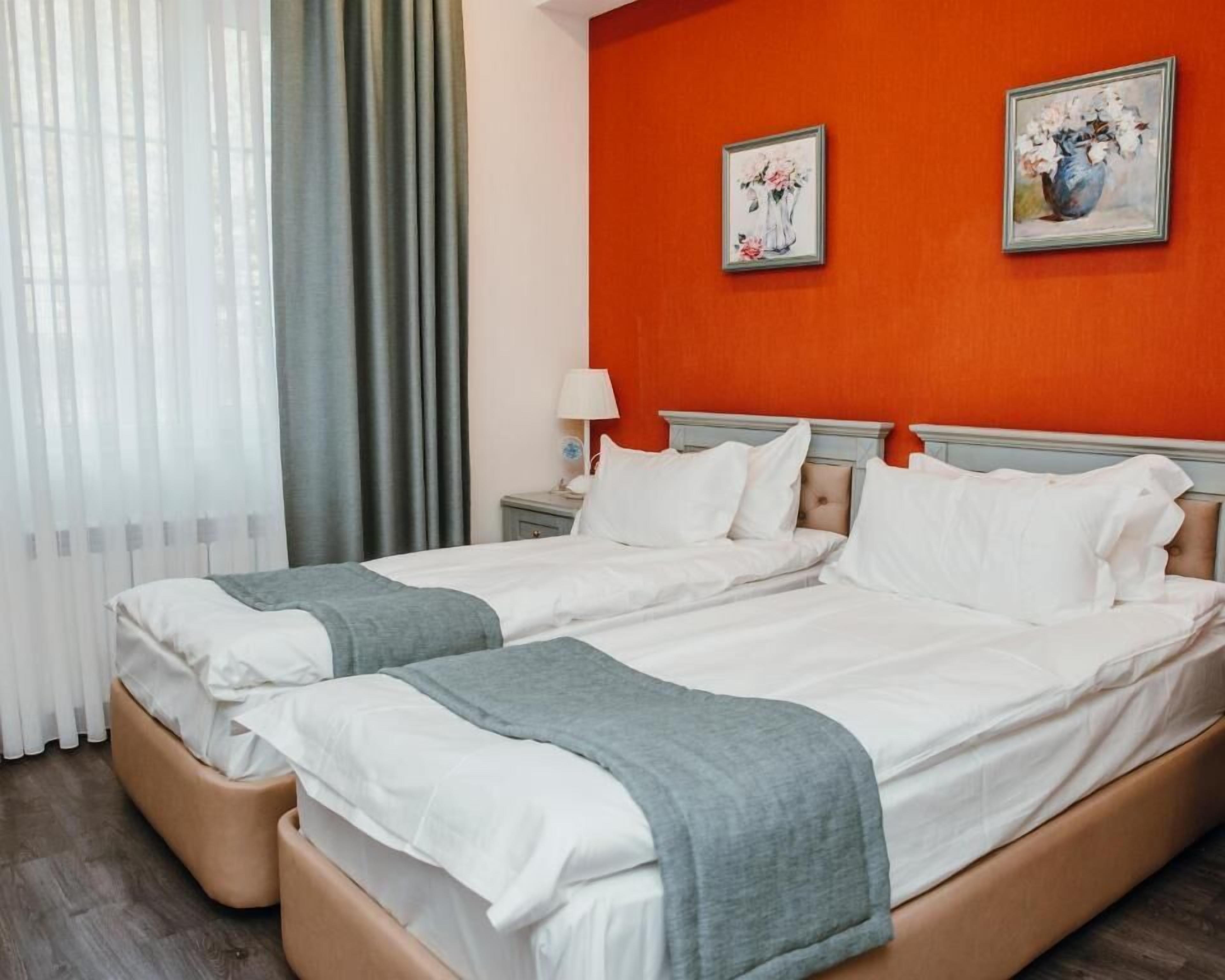A double bed with white linens and a grey throw, two bedside tables with lamps, a red accent wall, and two framed floral paintings.
