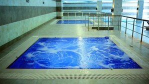 Uma grande piscina interior com um elemento aquático azul, rodeada por um chão em mosaico e um guarda-corpo de vidro.