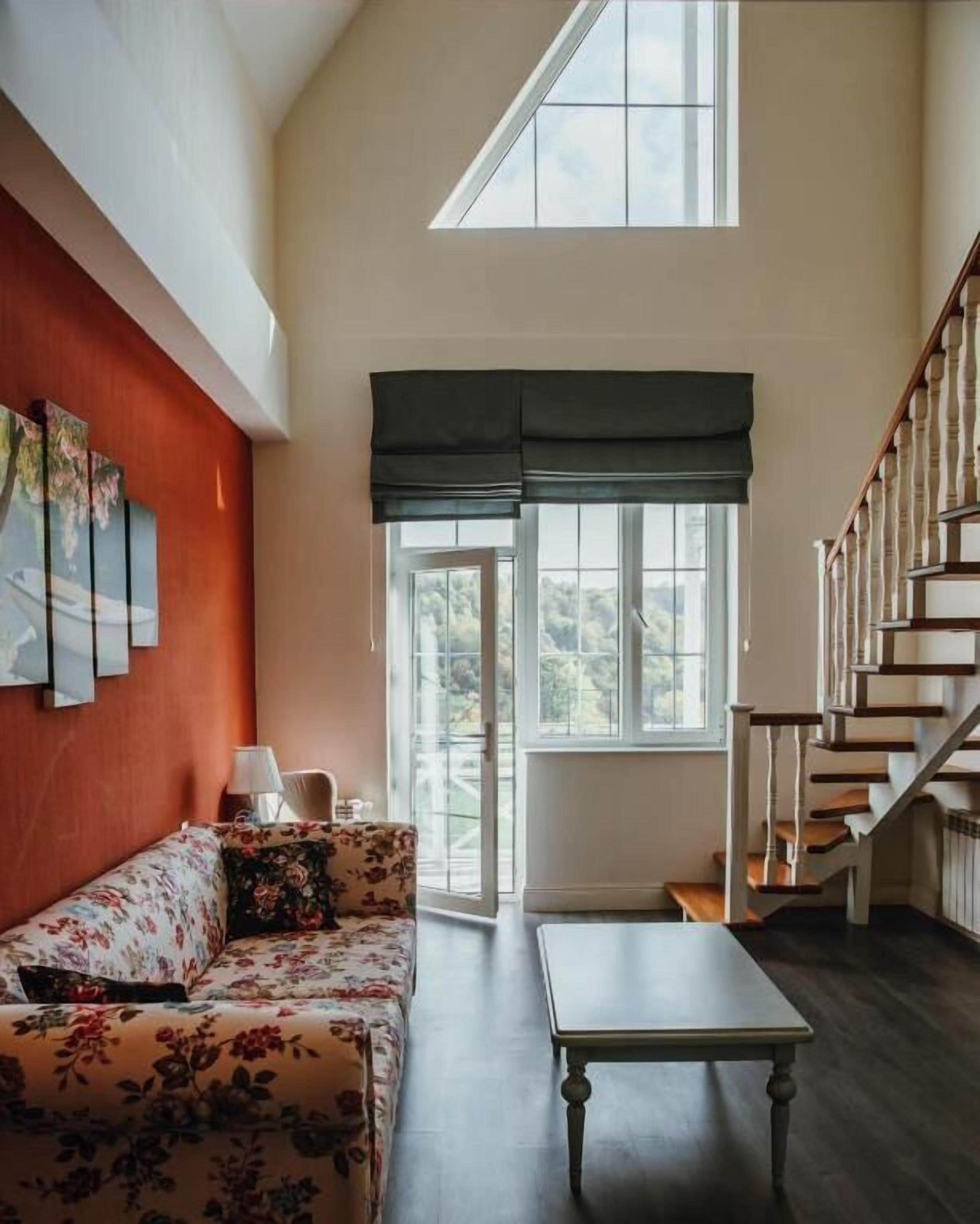 A spacious living room with a high ceiling, a wooden staircase, and a large window with a view of the outdoors.